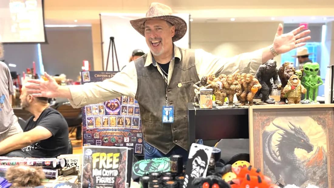 Professor B behind his convention table with both arms outstretched, surrounded by cryptid figures, plush creatures, illustrated cards, and a framed dragon painting.